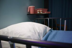 A close up of a hospital bed made up with a blue blanket and white pillow. Medications and pink plastic basins and a pink pitcher sit on a shelf in the background.