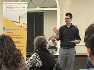 Doug Quattrochi, a man with dark hair wearing a black dress shirt and gray dress pants, gives a presentation in front of a room of people. A MassLandlords banner can be seen to the left.