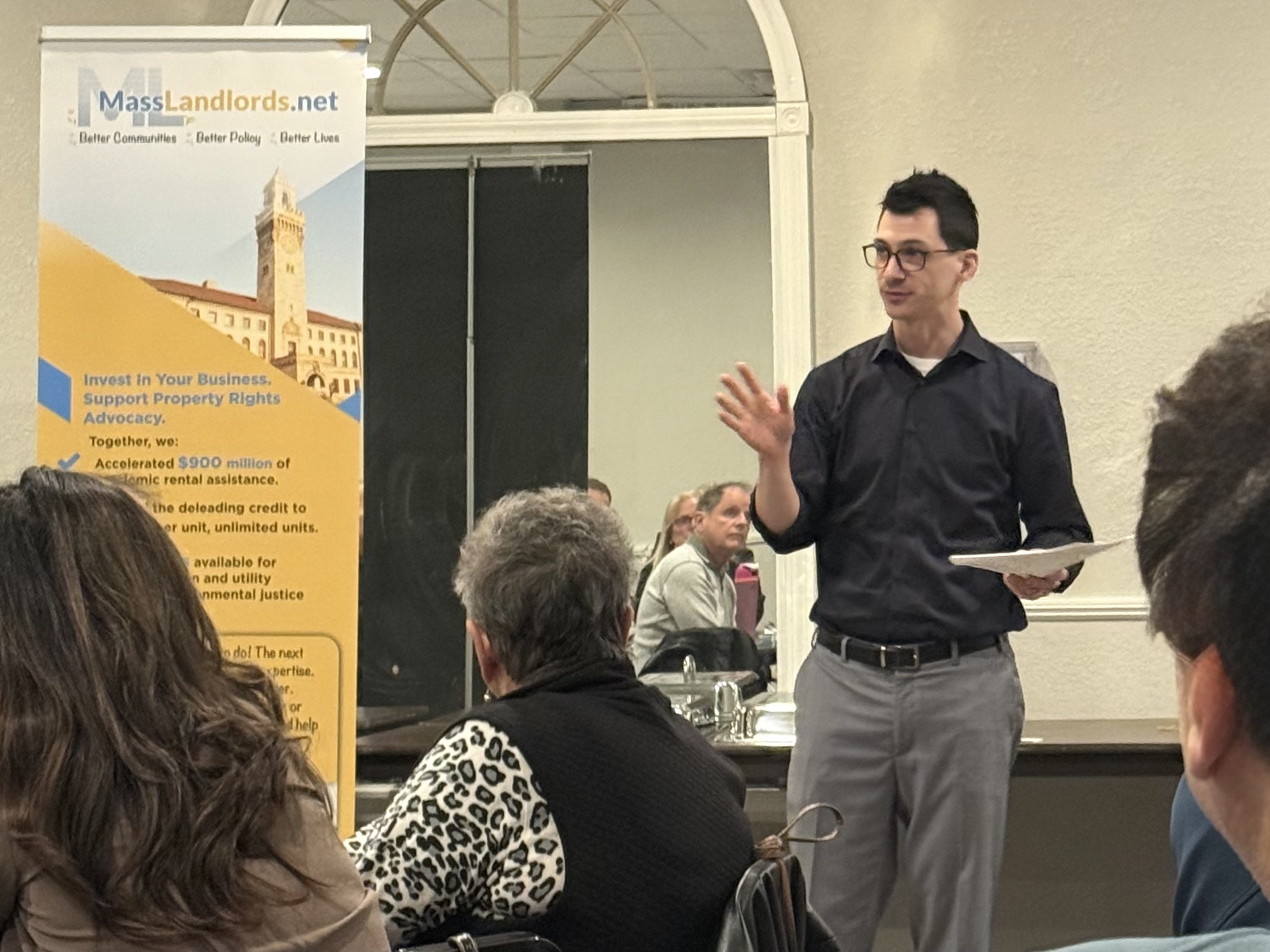 Doug Quattrochi, a man with dark hair wearing a black dress shirt and gray dress pants, gives a presentation in front of a room of people. A MassLandlords banner can be seen to the left.