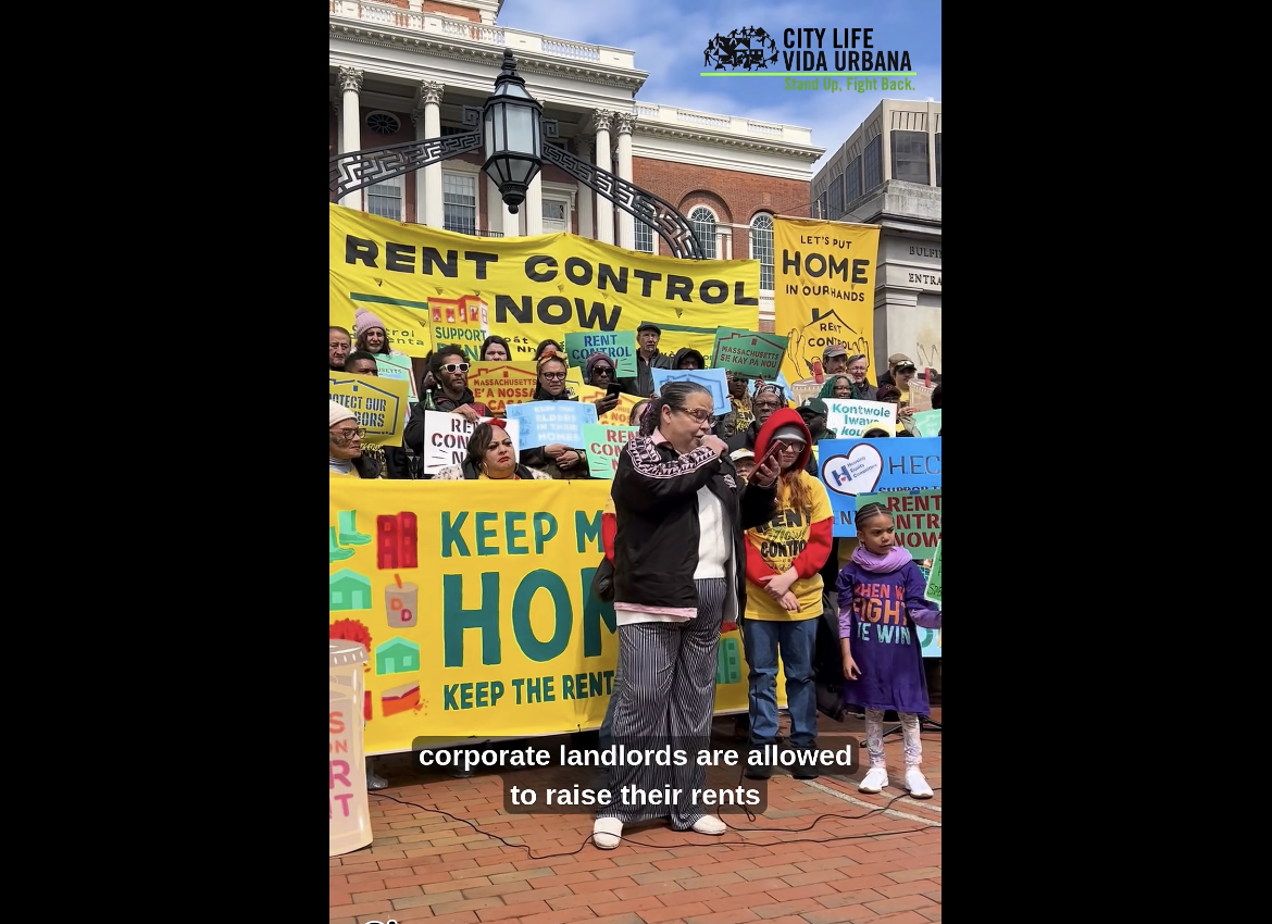 A screen capture with City Life/Vide Urbana’s watermark in the upper right corner shows pro rent control rally happening at the State House in Boston. A woman is speaking about rent control, the closed caption of this portion reads “corporate landlords are allowed to raise their rents.”