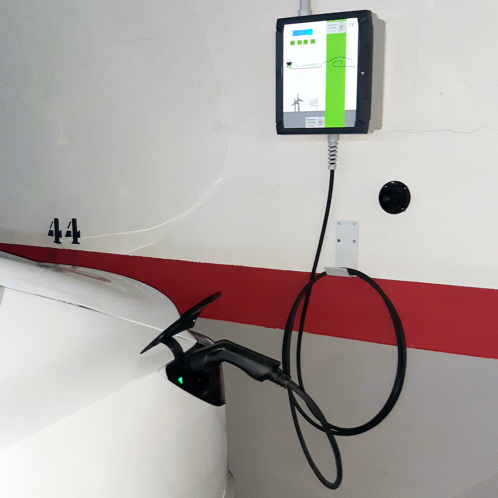 A car in a residential garage is connected to a charger cable, which is connected to a box on the wall, which is connected to a wire conduit. The charger is very small relative to the car and the garage.