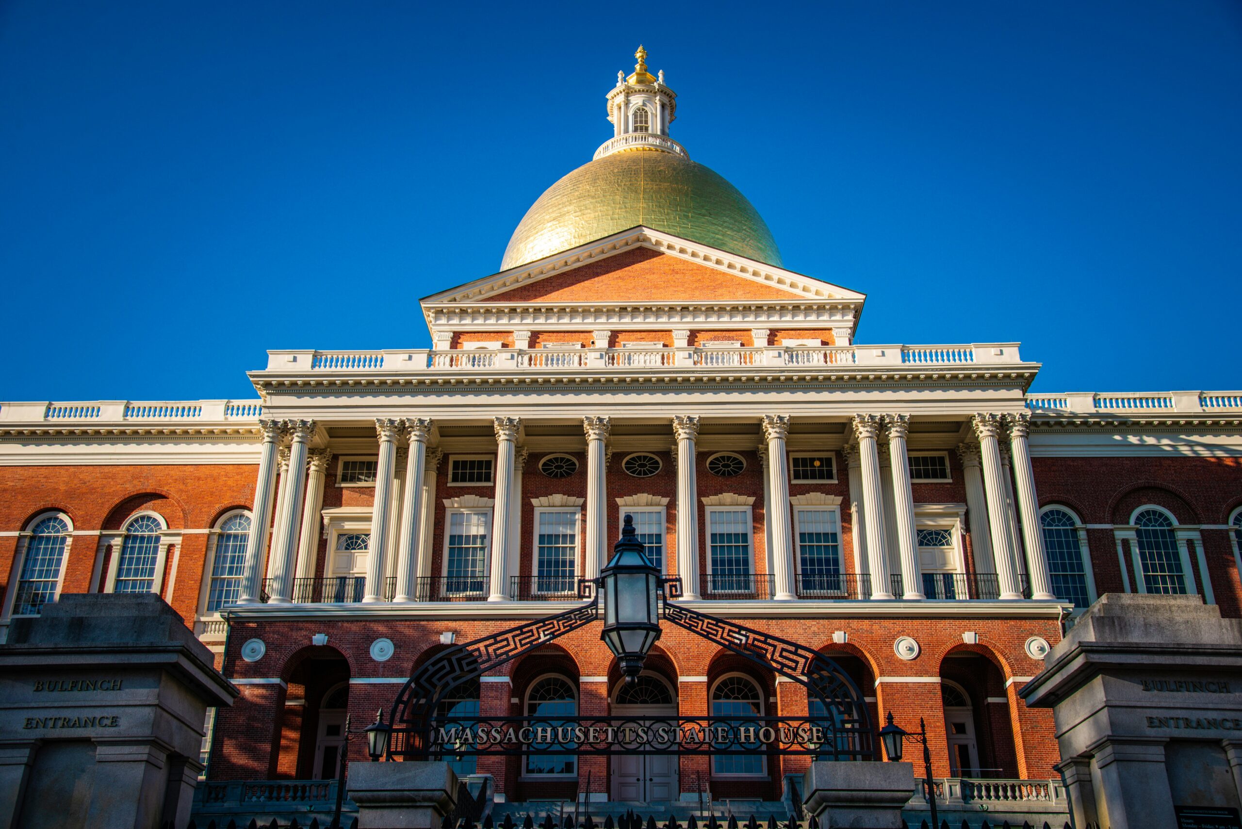 A close-up view of the Massachusetts State house with gold dome visible, and the front wrought iron gate that reads “Massachusetts State House” in gold block letters.