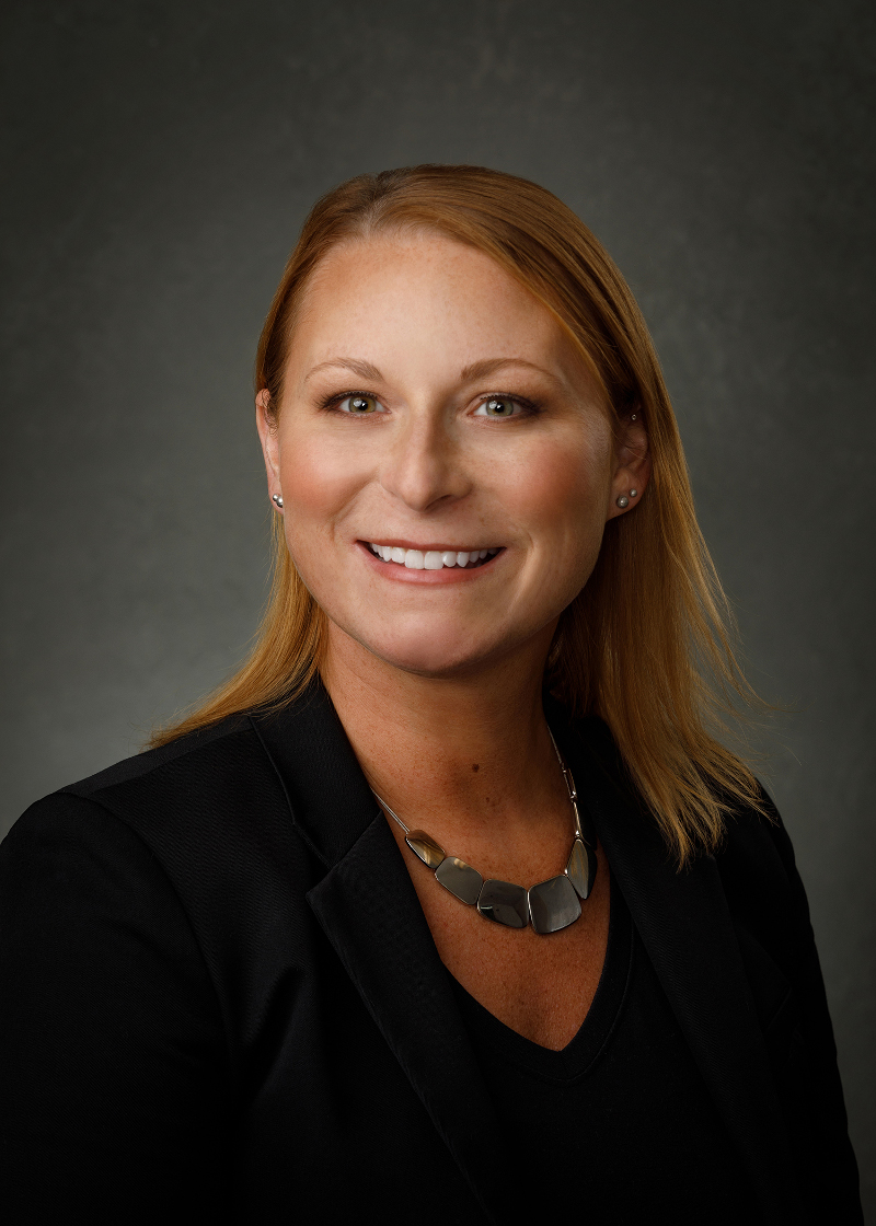 Attorney Melissa Gleick sits for a professional headshot with suit coat, smooth plate metal necklace, shoulder length hair, and subtly double- and triple-pierced ears. She smiles warmly and sincerely.
