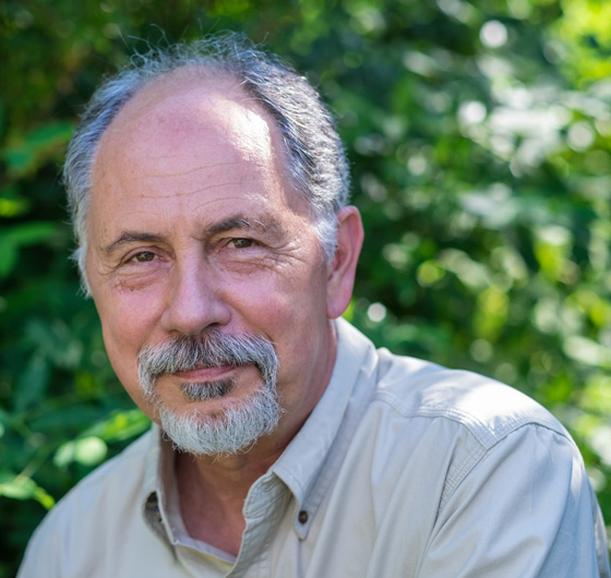 Tom Chase wears a goatee beard, close cropped hair and a button-down collared shirt open at the collar. He smiles warmly in front of a forest background.