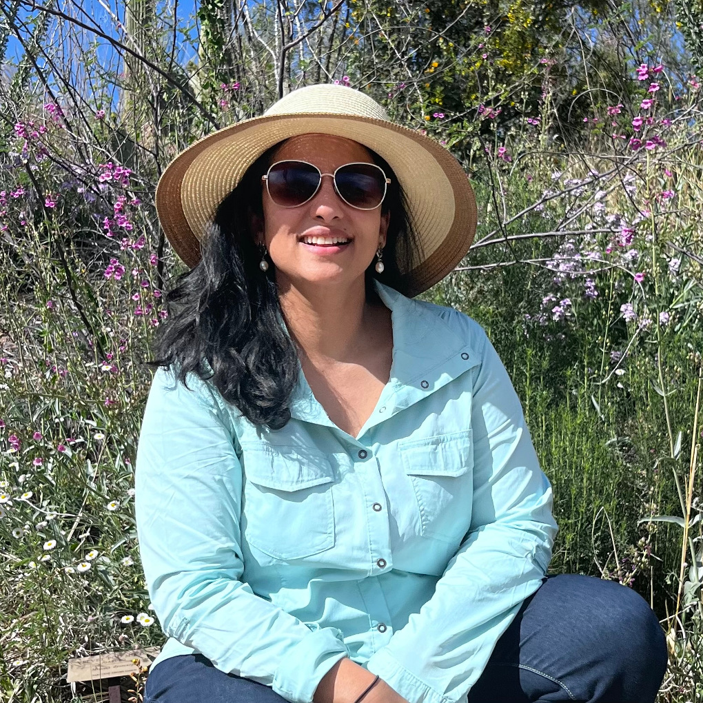 Tripti Travers-Thomas wears past-shoulder length hair, sunglasses, a neatly trimmed, broad brimmed straw hat, pearl drop earrings and a collared shirt. She smiles half laughing in front of a wildflower meadow.