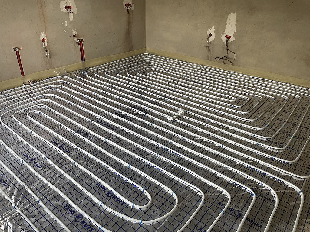 photo of a radiant floor pipe grid, with spiraling pipes meandering over a subfloor, with water supply pipes entering the room along a concrete wall.