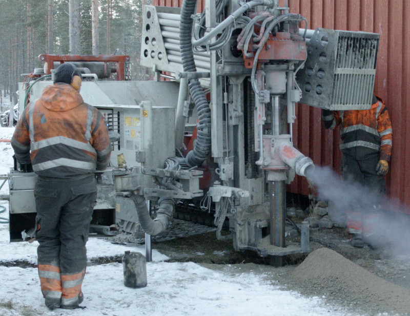 A man in dirty high-vis clothing operates a drill, which is spewing dust and soil to one side.