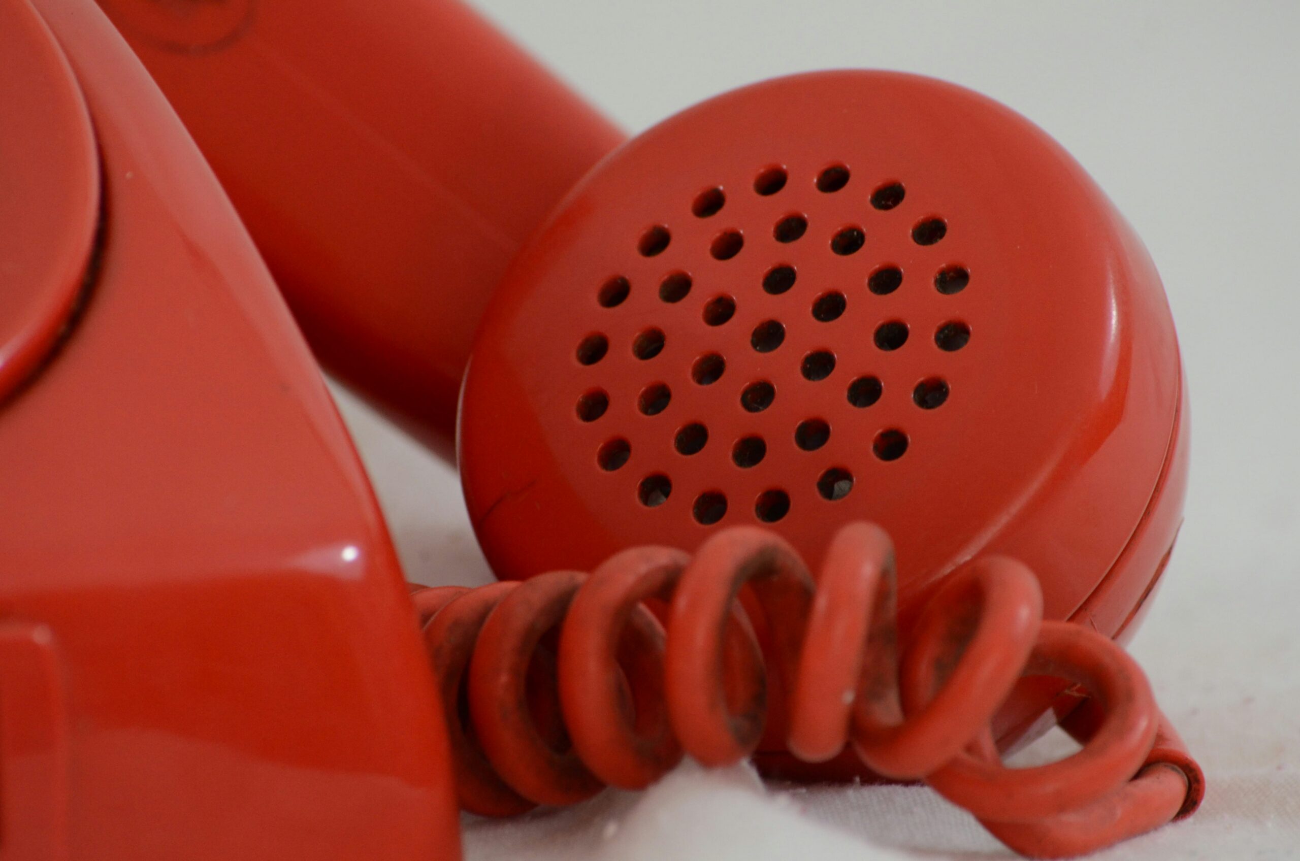 Alt – a close-up of the front corner of an old-fashioned red rotary telephone. Part of the coiled cord and receiver can be seen to the side.