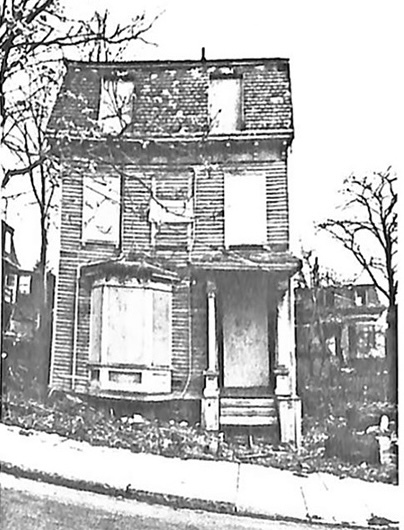 black and white, aged picture of a creepy dilapidated, two-story single family house with its door and windows boarded up and its exterior untended and trashy. A sidewalk runs in front of the house at a steep angle, and there are several trees around the house with no leaves. Trash and detritus litter the front and side yard.