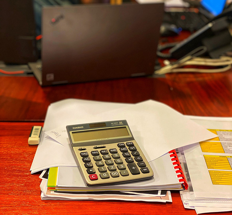 A stack of paperwork sits on a wooden desk. A calculator sits on top of the stack. A black laptop is open in the background.