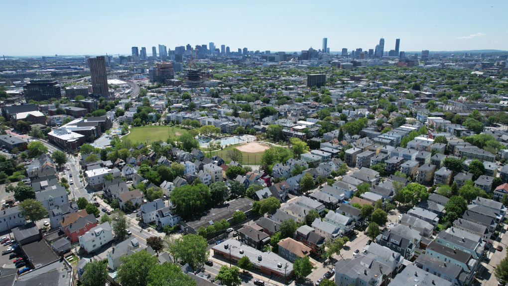 A bird's eye view of Somerville with the Union Square toward in the middle distance and the Cambridge and Boston skylines in the distance. There are three-deckers everywhere separated by roads and recreational fields.