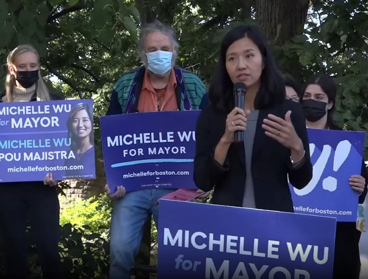 a photo of Boston Mayor Michelle Wu speaking at a campaign rally, with supporters in the background wearing masks and holding signs saying “Michelle Wu for Mayor” in English and Haitian Creole.