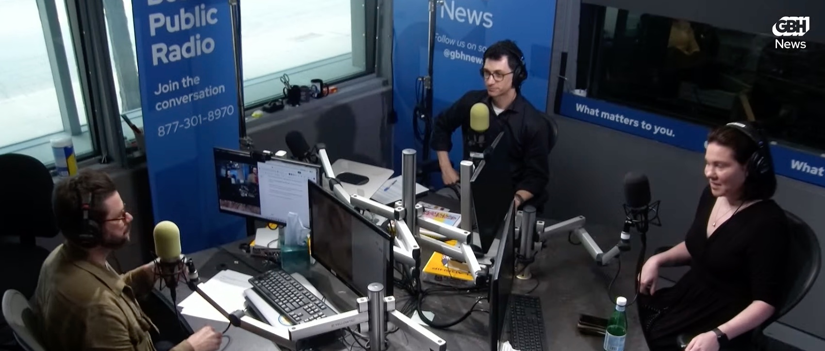 screen shot of a GBH News panel, in which panelists MassLandlords Executive Director Doug Quattrochi and Jennifer Smith, a reporter with Commonwealth Beacon, are seated across from radio host Edgar B. Herwick III, all wearing headphones and facing each other, with desks in front of them cluttered with computer monitors, microphones and mic stands, a bottle of water, and two windows in the background onto the street. Placards on the background wall read “Public Radio, Join the conversation,” and “What matters to you.”
