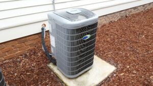 A close up photo of a heat pump exterior unit sitting in mulch next to the outside wall of a house, showing part of a brick foundation and house siding.