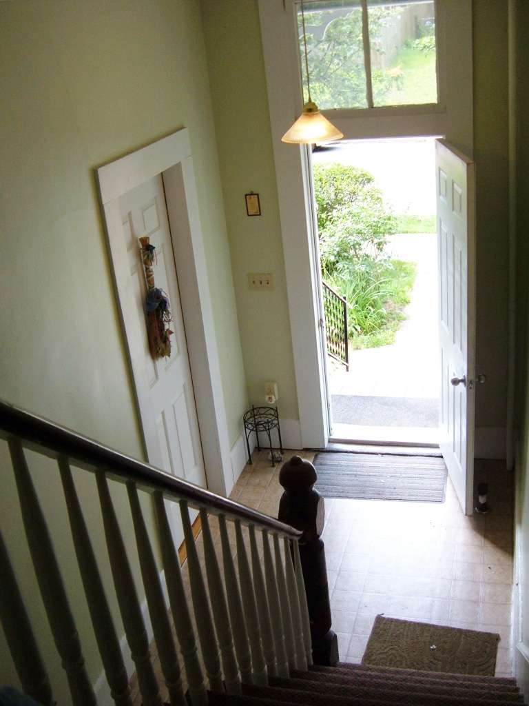 photo of an interior front hallway of a multifamily building looking down from a stairway out an open front door onto a sunny stoop and front sidewalk.