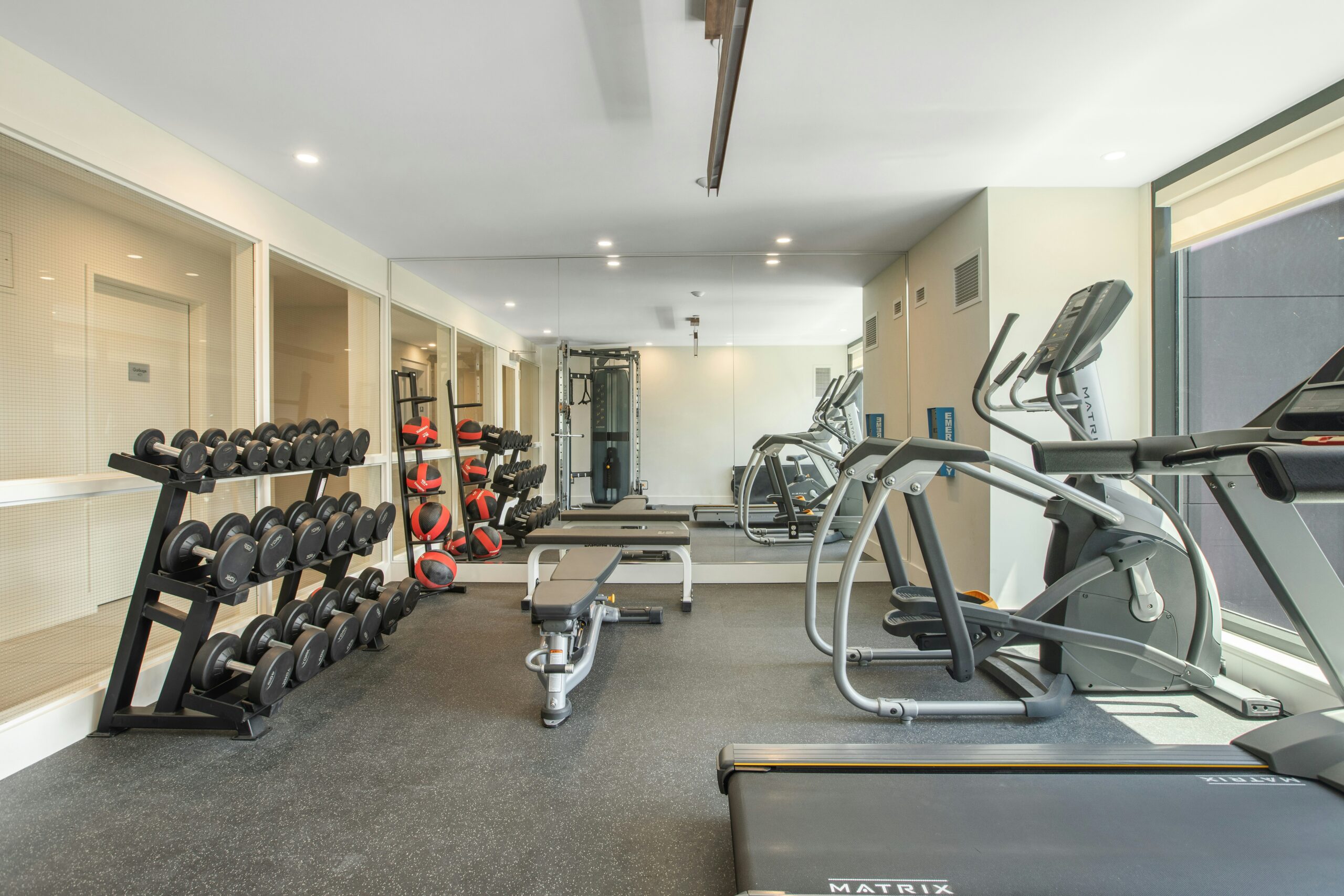 Photo of a small gym, with free weights along one glass wall, medicine balls, weight benches and treadmills and elliptical machines lining the opposite wall of a long room with gray floor.