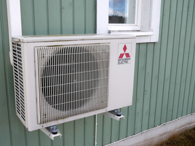 An electric Mitsubishi heat pump is installed under a window of a house with green siding.