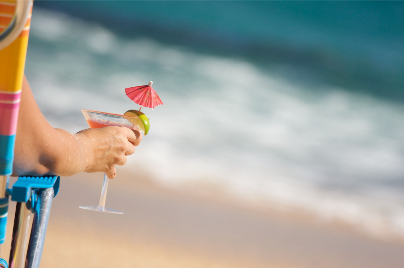 photo of a person sitting in a beach chair holding a pink cocktail in a martini glass with a lime wedged on the rim and a tiny pink umbrella. A blurred beach and ocean in the background.