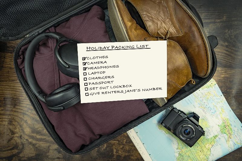 Photo of an open valise packed with walking shoes, headphones, and on top, a piece of paper labeled “Holiday Packing List,” with items listed such as clothes, camera, headphones, laptop and passport. Beside the valise on a table is a map and a camera.