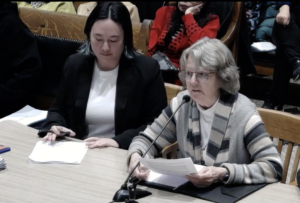 Two women sit at a table in the State House. The woman on the left is looking down at some papers. The woman on the right is speaking into a tabletop microphone.