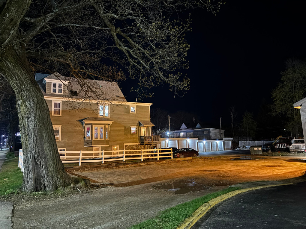 The scene covers from left to right a public road, a tree, a residential building, a garage, a business parking lot and the edge of a business building. It is night, but the scene is as bright as day with light pollution coming from all directions and plastering the entire side of the residential building.