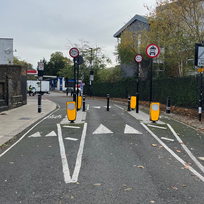 An intersection has a bike lane running on either side of a road. A single lane for vehicle traffic is possible, but large metal bollards stick up from the road blocking the intersection to cars. Bikes can pass the bollards to either side.