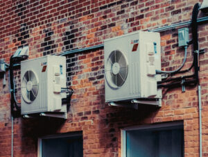 two external heat pumps are affixed to the side of a brick building above the windows.