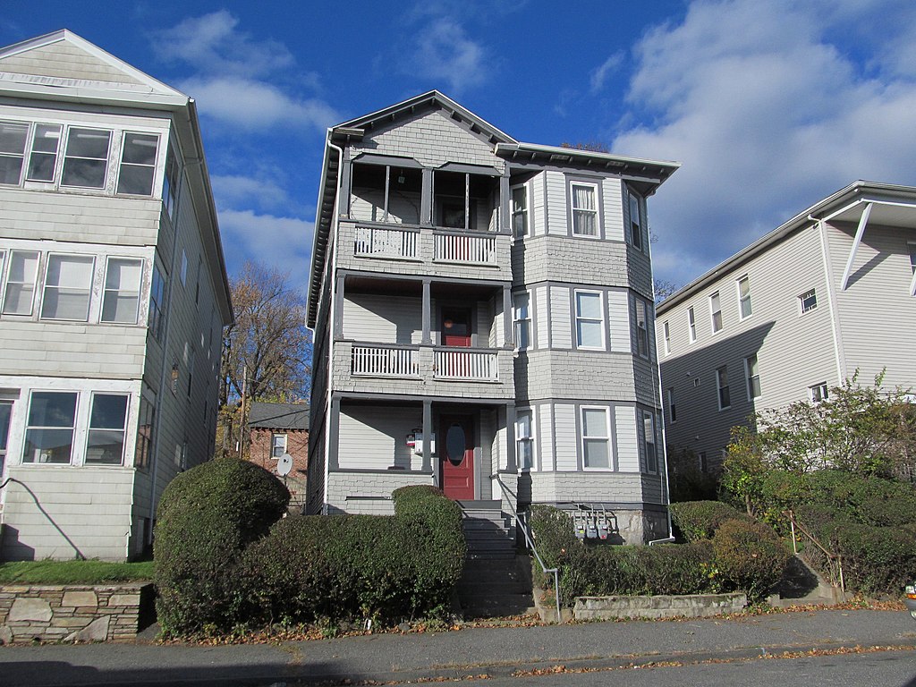 photo of three triple decker multifamily buildings, each painted gray, the middle one landscaped with shrubs around the front stoop, with blue sky and some wispy clouds overhead.