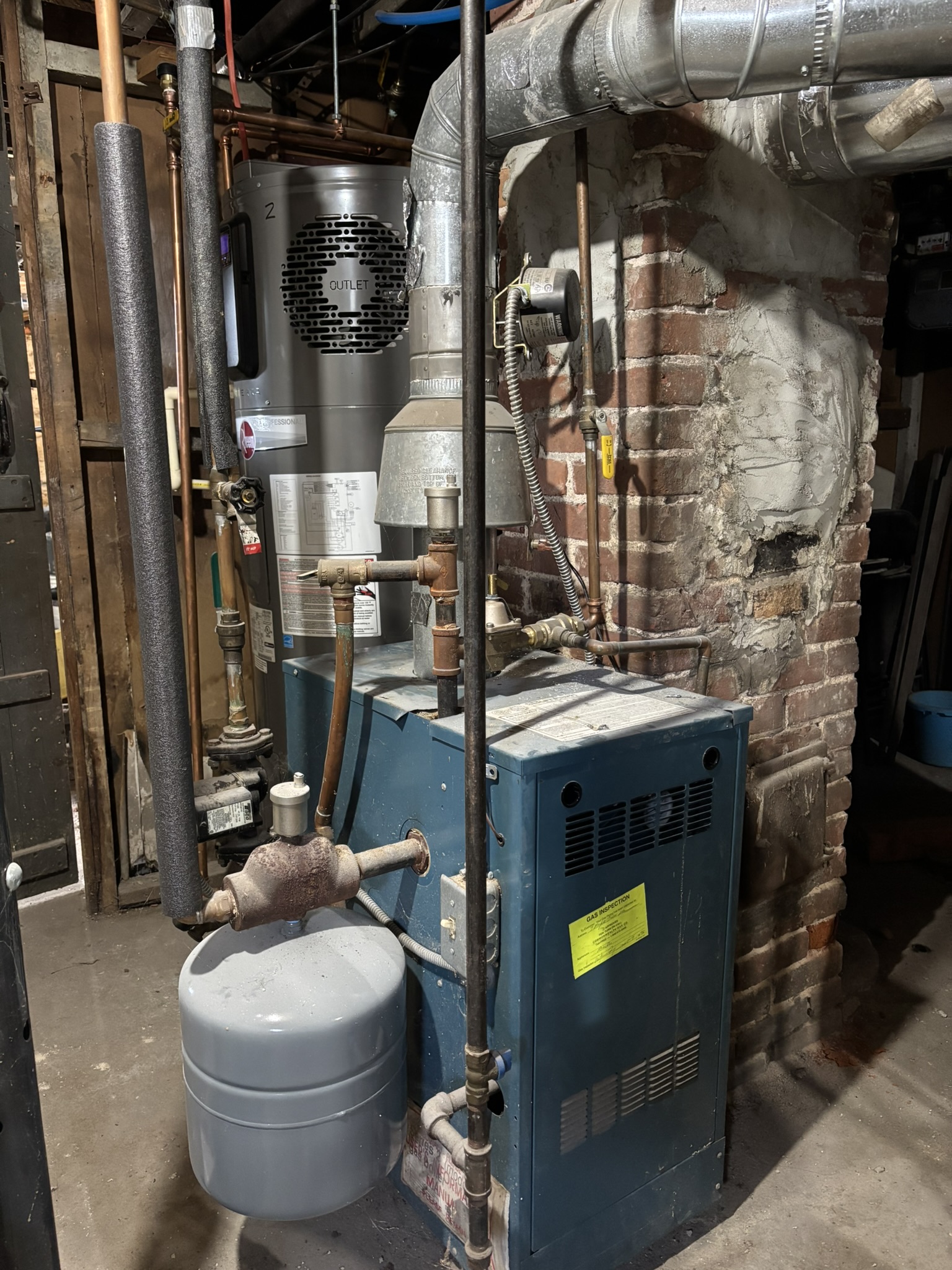 A blue boiler with expansion tank sits beside a brick chimney. The boiler is old and dusty.