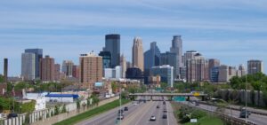 A panoramic photo of downtown Minneapolis, Minn., viewed on approach from the south, with a cluster of new and old high rise business buildings and apartments, a four-lane highway leading to the city, and a blue sky backdrop with slight, wispy clouds.