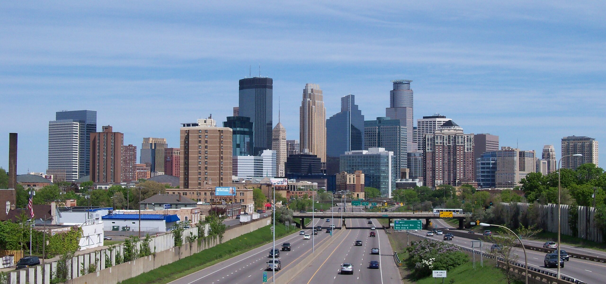 A panoramic photo of downtown Minneapolis, Minn., viewed on approach from the south, with a cluster of new and old high rise business buildings and apartments, a four-lane highway leading to the city, and a blue sky backdrop with slight, wispy clouds.