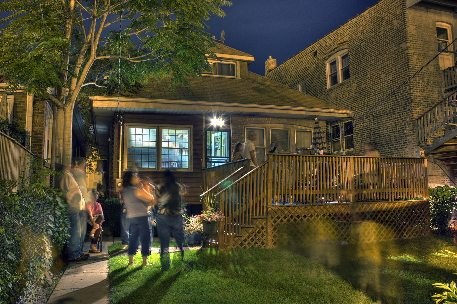 photo of the backyard of a small house built very closely between a neighboring house and a brick multifamily building, with blurred images of people gathering, mothers holding babies in the grass, with a large deck extending off the back of the house, a tree overhead, and a dark blue evening sky in the background.