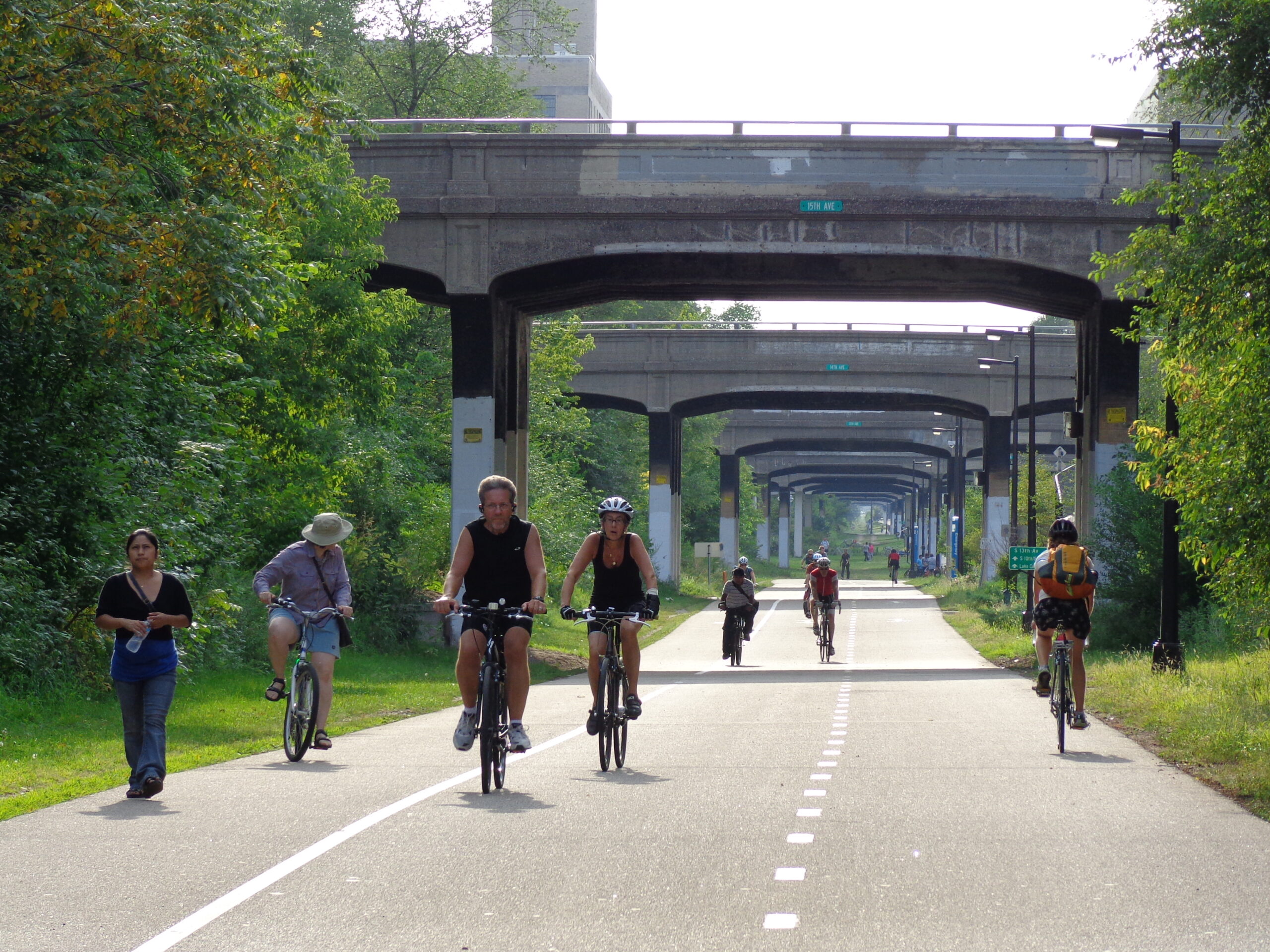A photo of the Minneapolis Midtown Greenway, part of the city’s network of bicycle and pedestrian pathways, showing a concrete, two-lane bike road with white dotted dividing line down the center and a solid line on the side defining the walking path, with numerous people on bikes coming toward and riding away from the camera, with a series of underpasses leading into the distance.