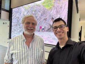 A photograph of two Caucasian men standing side by side in a building lobby with a large projector screen in the background. The man on the left has light hair and a beard and wears a light-colored striped button-down shirt with two buttons undone. The man on the left has dark hair and wears a black polo shirt. Both are smiling.