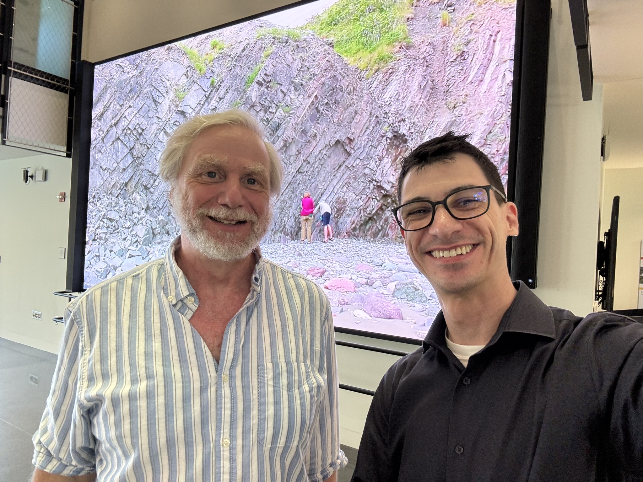 A photograph of two Caucasian men standing side by side in a building lobby with a large projector screen in the background. The man on the left has light hair and a beard and wears a light-colored striped button-down shirt with two buttons undone. The man on the left has dark hair and wears a black polo shirt. Both are smiling.
