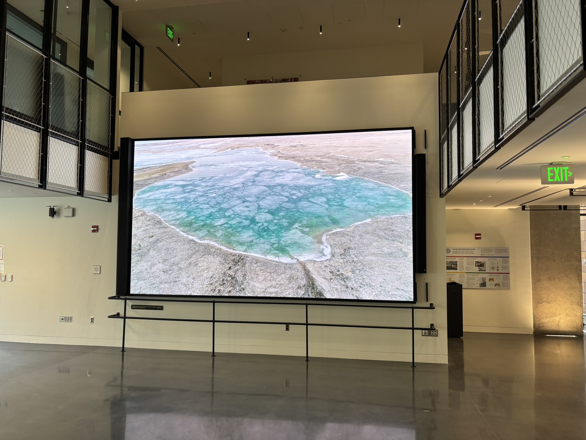 A photograph of a large lobby space with high ceilings and shiny floors. A large projection screen shows a multicolored natural image of blues, greens and browns.