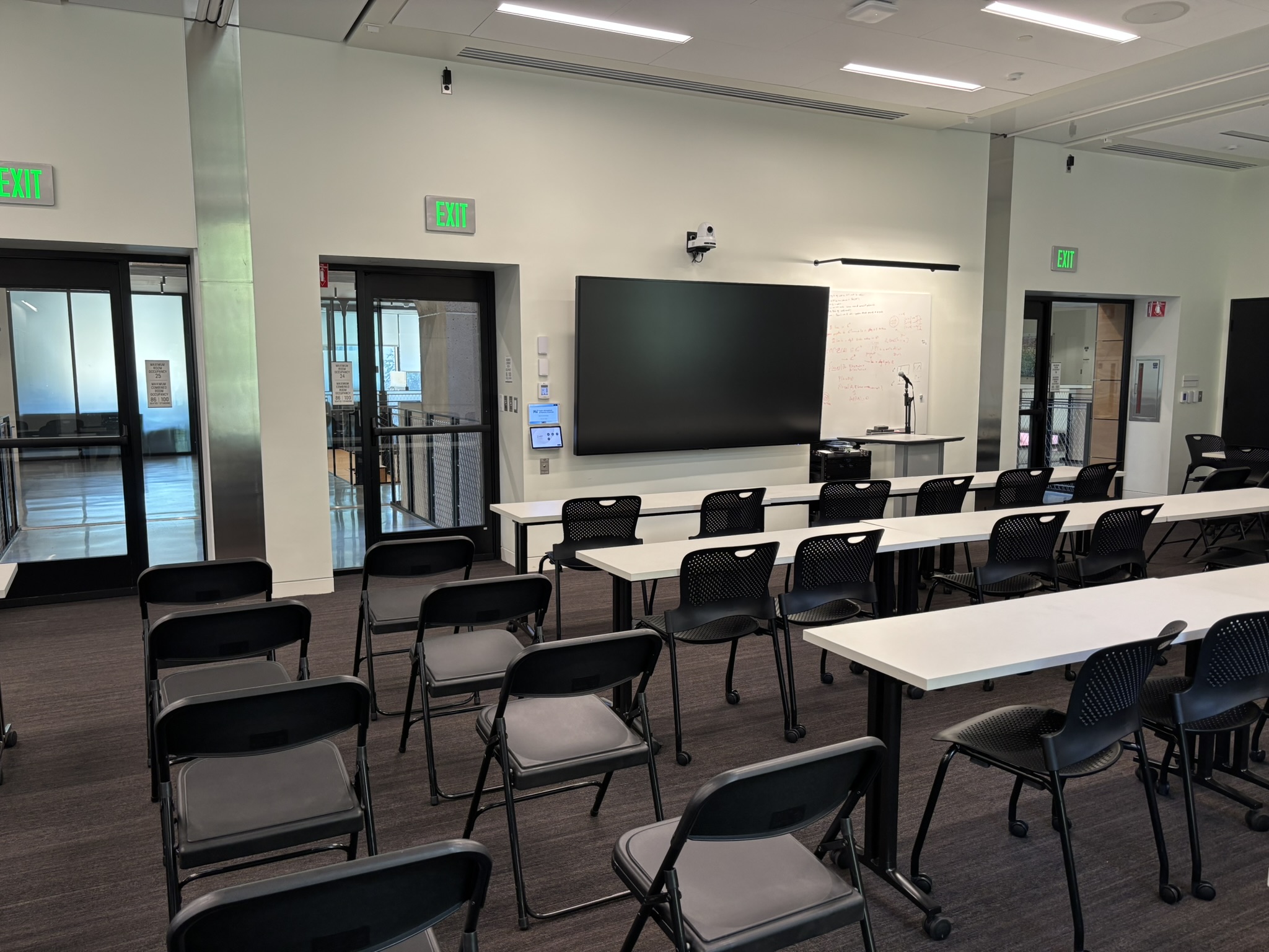 a photograph of an empty classroom with three long white tables. Black chairs are lined up at each table, with overflow chairs to the left in rows.