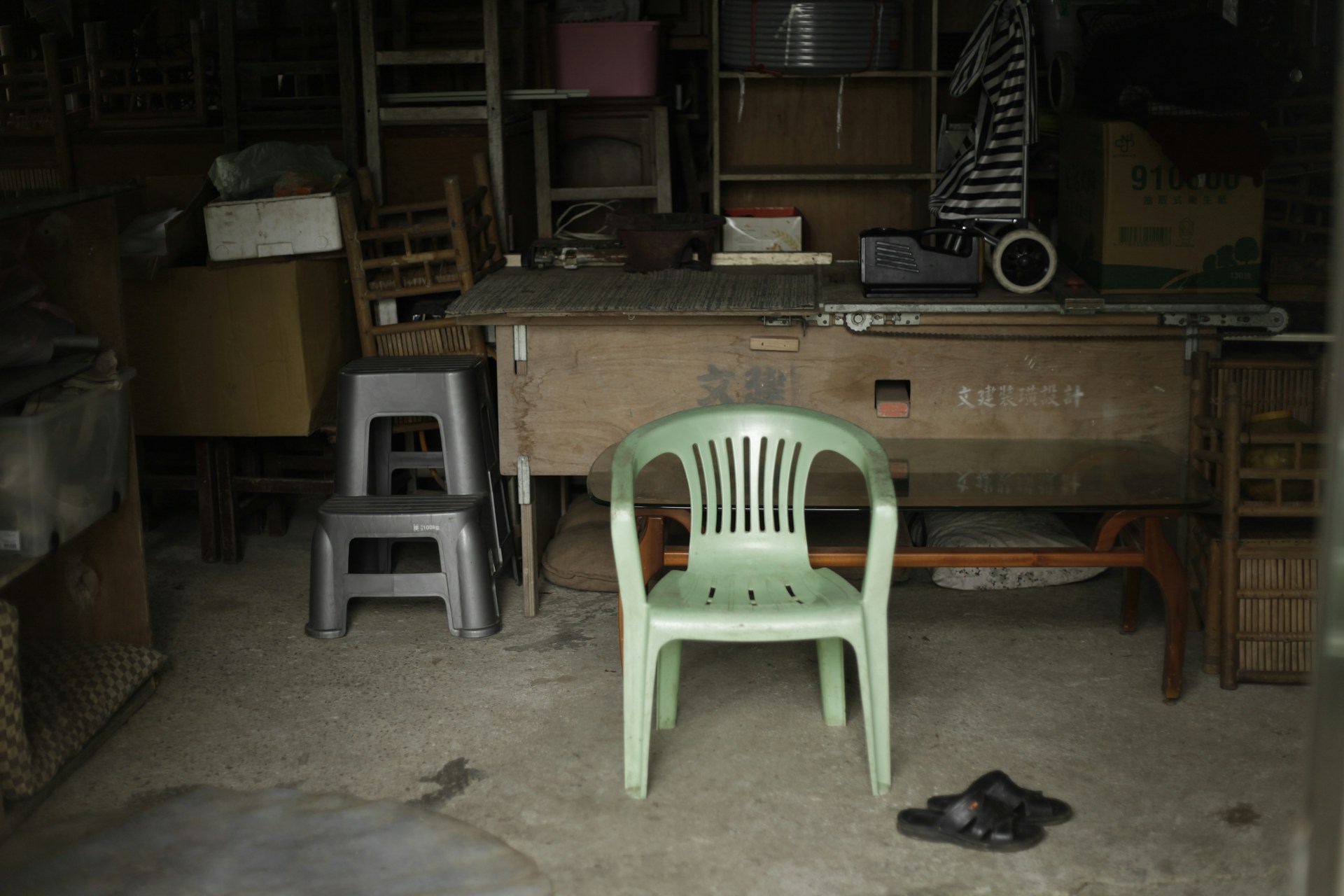 A residential garage space is full of old furniture and some tools and other items. A green lawn chair sits front and center in front of an old desk or workbench.