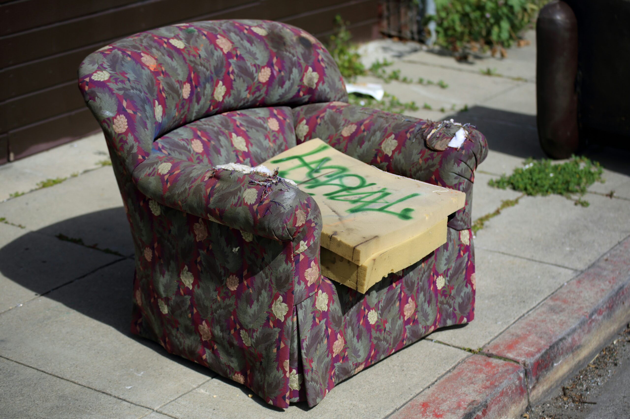 A dark pink floral armchair sits on a sidewalk curb. The seat cushion cover has been ripped off, exposing the foam, which someone has put a spray painted with graffiti.