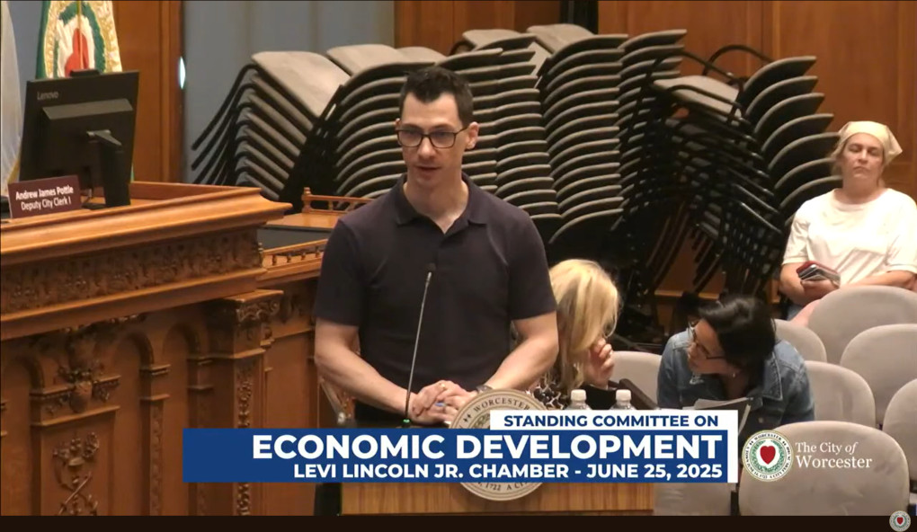 A view of the Levi Lincoln Jr. chamber at Worcester City Hall shows wood paneling behind stacks of unused chairs in the background. In the foreground Doug Quattrochi wears a collared, short-sleeve polo shirt and glasses. Three people are visible in the audience, one of whom may be paying attention. The other two are talking. Quattrochi addresses councilors off-camera.