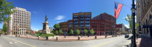 panoramic photo of downtown Portland’s Monument Square, featuring a brick-palavered park with statues amid several red-brick and modern office buildings, with city streets running down both sides of the park against a bright blue sky.