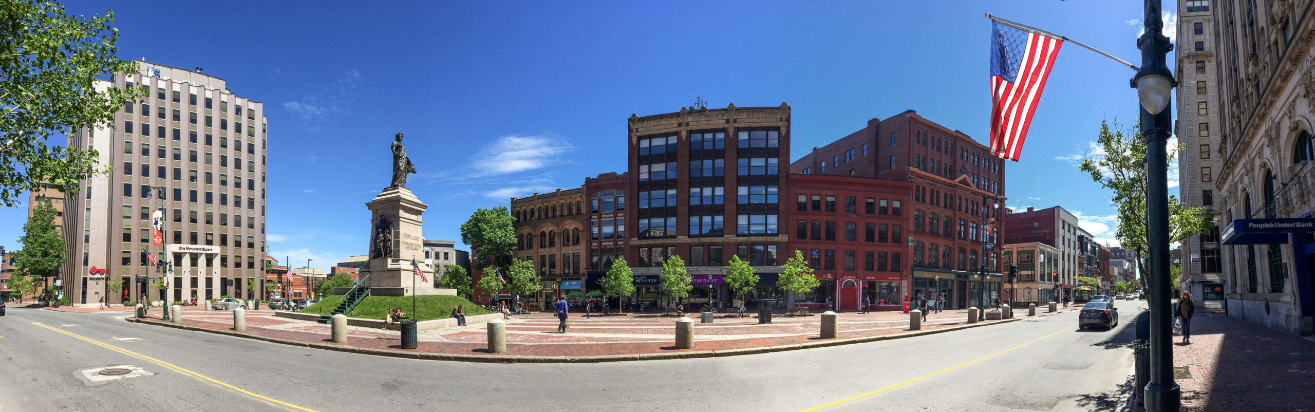panoramic photo of downtown Portland’s Monument Square, featuring a brick-palavered park with statues amid several red-brick and modern office buildings, with city streets running down both sides of the park against a bright blue sky.