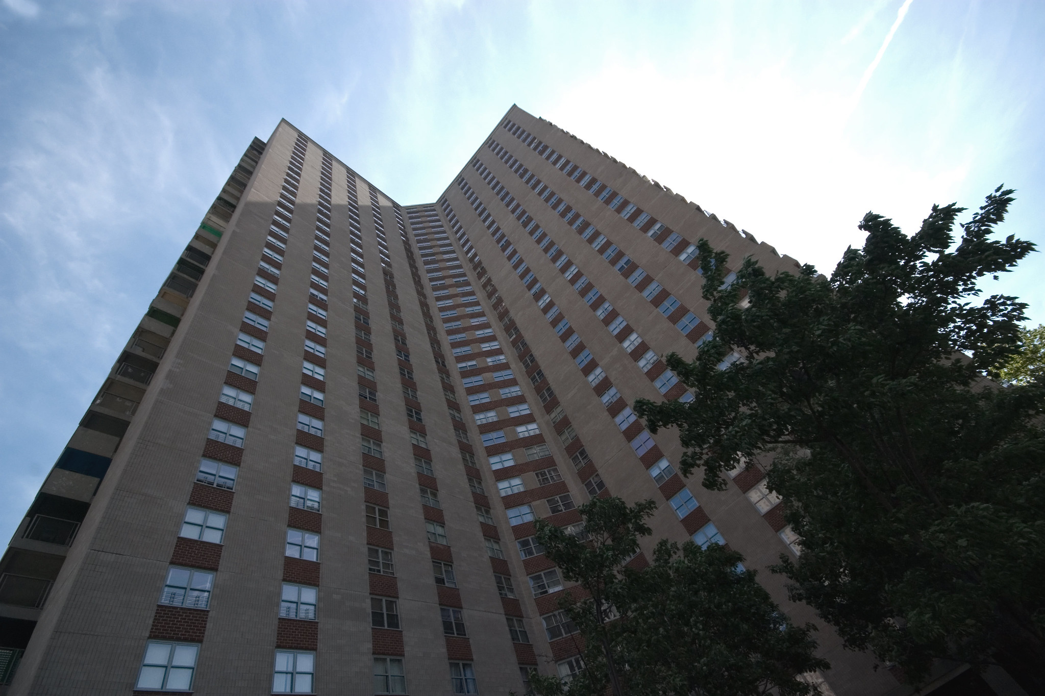 photo of a modern skyscraper apartment building, from the ground looking up its floors, toward a blue sky with wispy clouds high above, and a couple trees in the foreground.