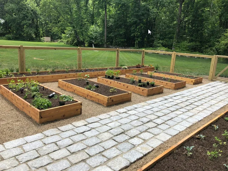 Cobblestones mark a path among low wooden boxes with open tops. In the boxes is dark soil with plants being planted in them. A smaller yard is visible in the distance.