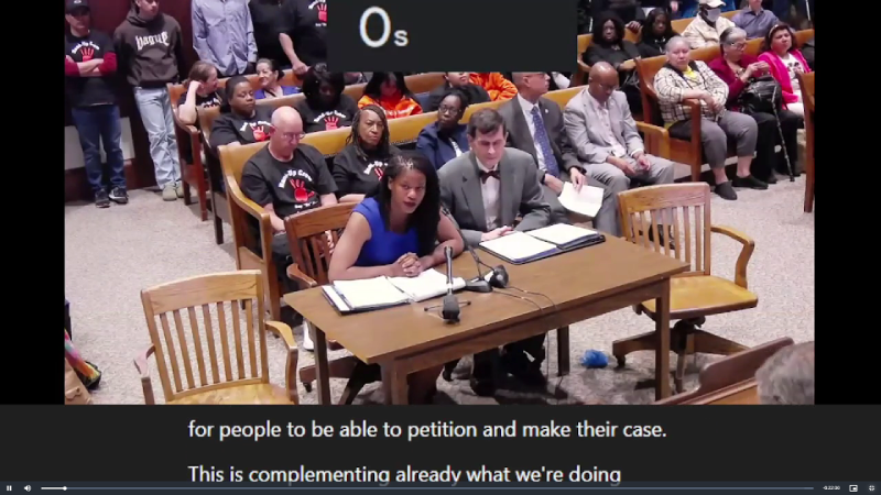 : A view from the State Housing hearing camera looks down on a packed hearing room with thin commercial carpet, wood table and four chairs for testimony. Two chairs are occupied, at left by Senator Edwards in a sleeveless dress and at right by Greg Vasil in a suit with bow tie. Behind them are four rows of benches full of people waiting to speak and a line of people standing at the back of the room.