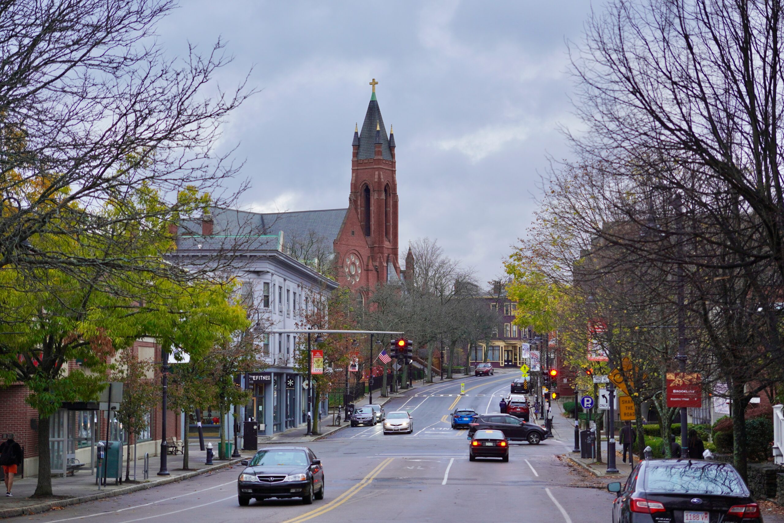 A photograph of a tree-lined street in Brookline, with several cars on the road and a church in the distance.