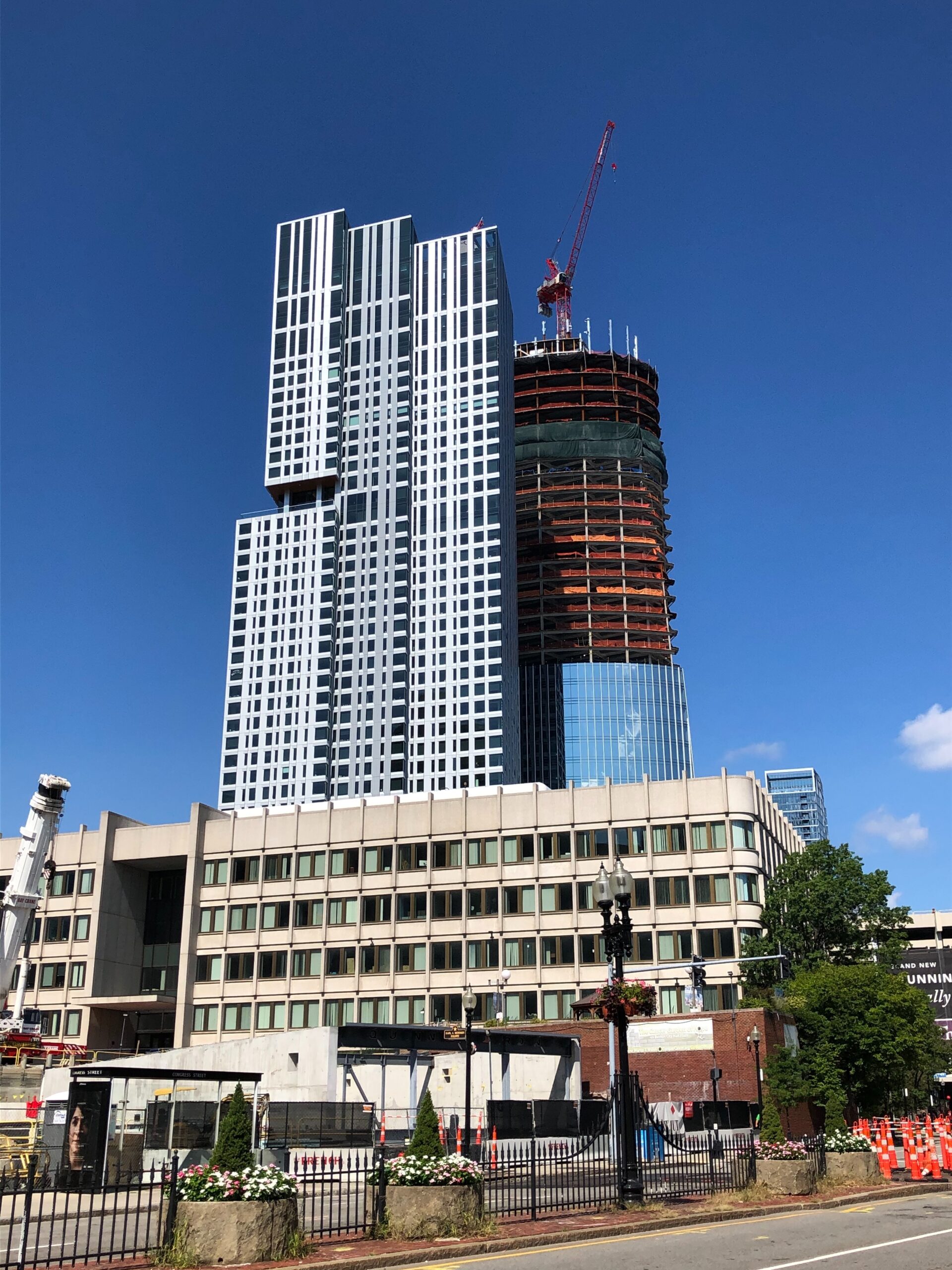 a photo of The Sudbury, a high rise Boston apartment building that came online in 2020, during its construction, with an incomplete tower with a crane on top, and an older concrete building in the foreground.