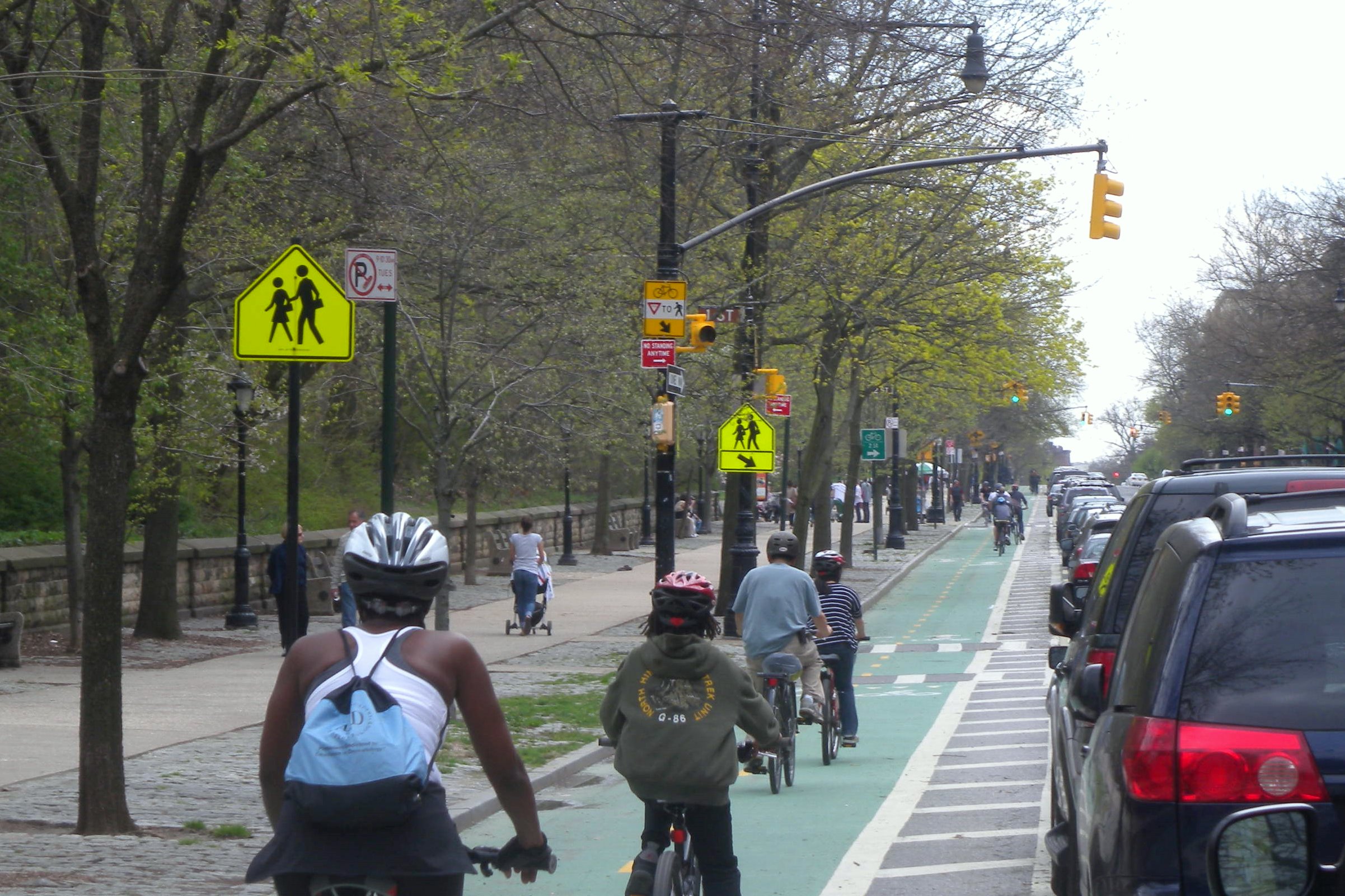 phot of a green-painted bicycle lane running along the side of a Boston street, with numerous cyclists in the lane and cars parked on the street, a wide sidewalk on the other side with many pedestrians, and a stone wall bordering a park, and trees starting to bloom. 