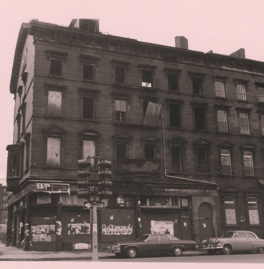 photo of a large, four-story, dilapidated building on a corner in South Boston in the 1970s, with cars from that era parked along the street. Several windows are missing, some are boarded over, graffiti painted on others, old flyers taped on boards at street level, very run down.