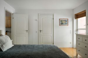 A photograph of a bedroom with the door open, revealing a kitchen in the next room. A double bed with a gray bedspread takes up most of the room, with a dresser and window on the opposite wall. The walls are white.